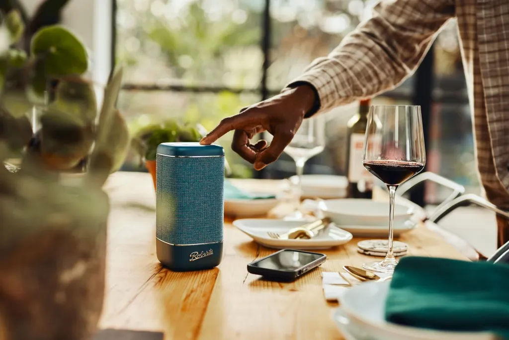 Een hand bedient een blauwe Roberts Beacon-speaker op een gedekte tafel.