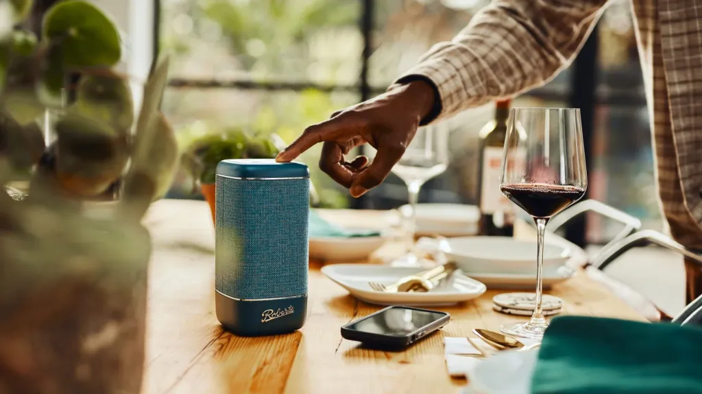 Een hand bedient een blauwe Roberts Beacon-speaker op een gedekte tafel.