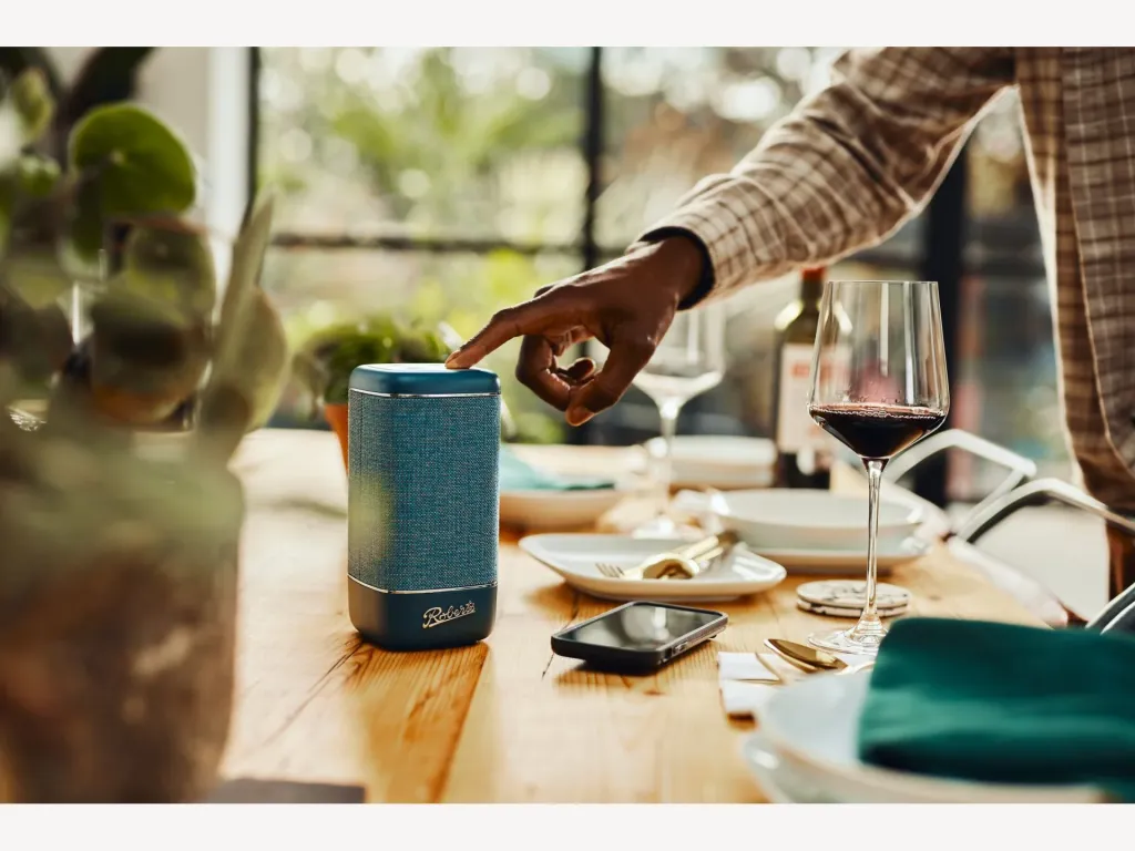Een hand bedient een blauwe Roberts Beacon-speaker op een gedekte tafel.